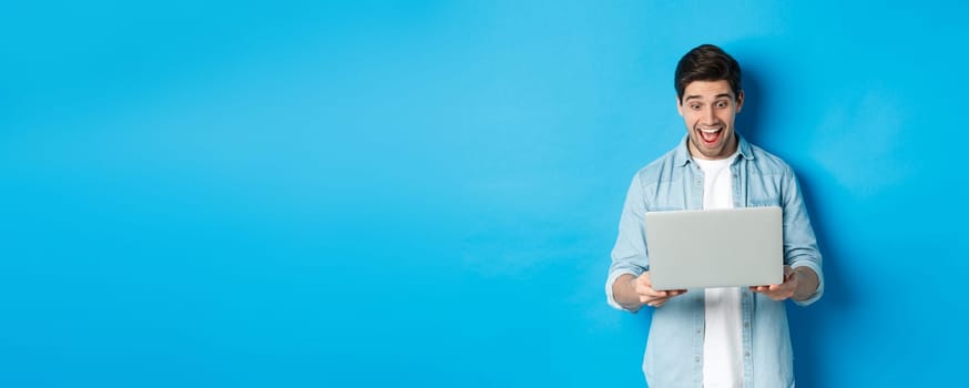 Image of amazed and happy man reacting to special offer in internet, looking at laptop excited, standing against blue background.