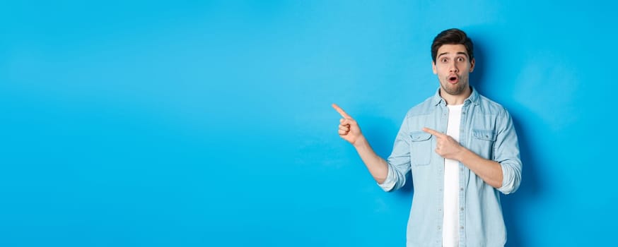 Portrait of surprised adult man in casual clothes showing announcement, pointing fingers left and looking amazed, standing against blue background.