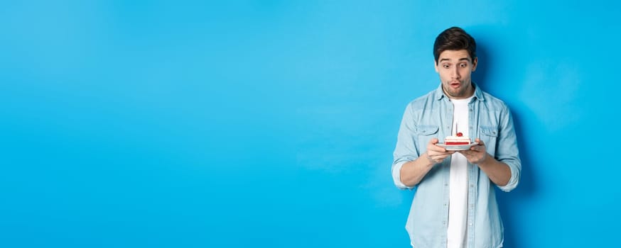 Birthday. Surprised man looking at b-day cake, standing against blue background.