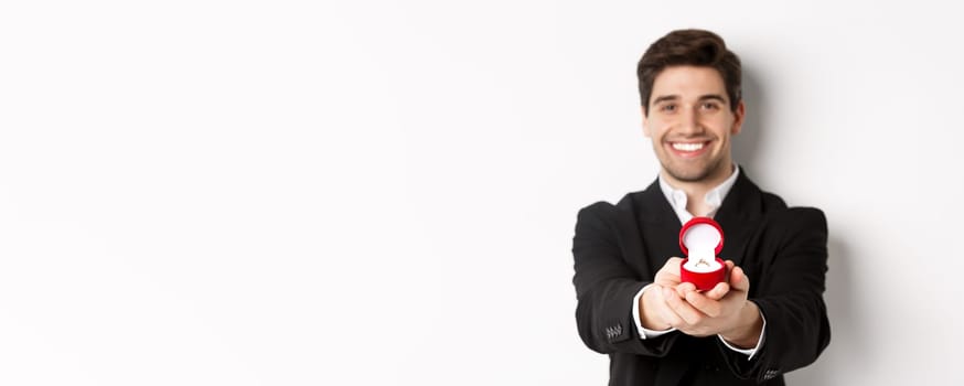 Image of handsome man looking romantic, giving you an engagement ring, making proposal to marry him, standing against white background.