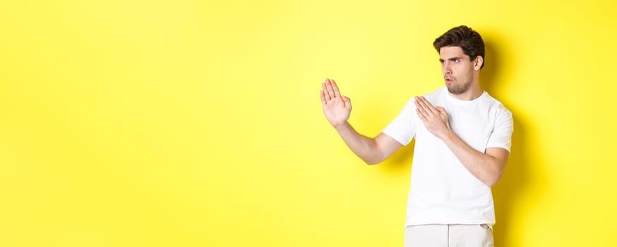 Man showing kung-fu skills, martial arts ninja movement, standing in white t-shirt ready to fight, standing over yellow background.