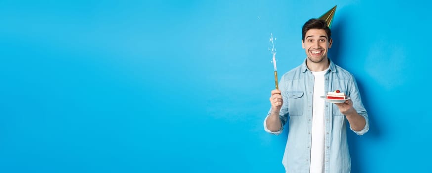 Happy young man celebrating birthday in party hat, holding b-day cake and smiling, standing over blue background.