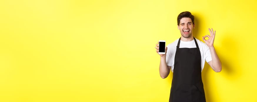 Handsome coffee shop worker showing ok sign and smartphone screen, recommending application, standing over yellow background.