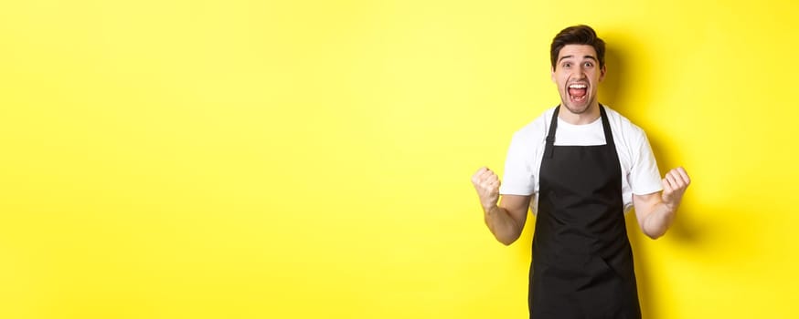 Excited coffee shop owner in black apron celebrating, making fist pump and shouting for joy, achieve goal, standing against yellow background.