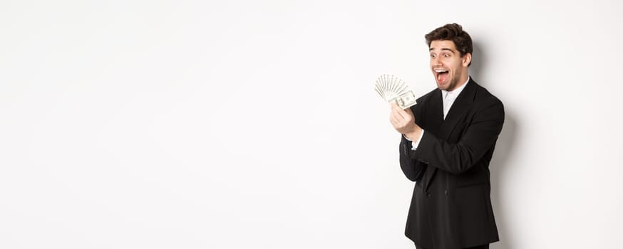 Image of excited handsome businessman in suit, looking amazed at money, winning cash, standing over white background.