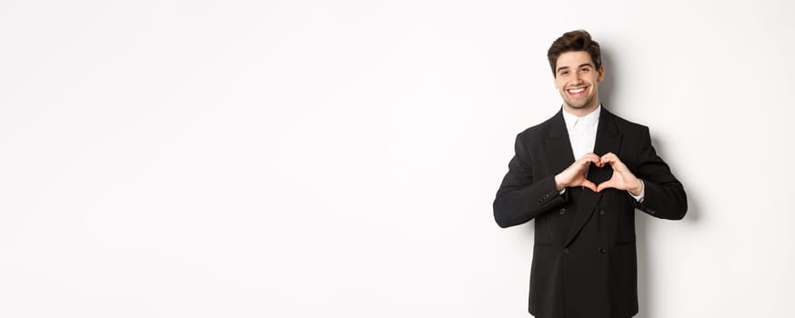 Image of handsome groom in black suit, showing heart sign and smiling, express love and sympathy, standing over white background.