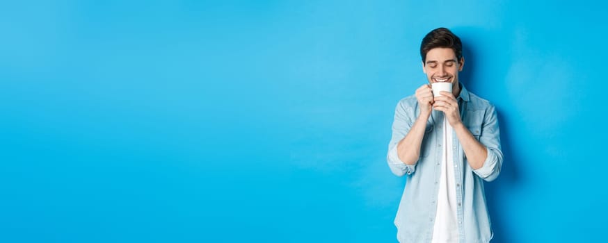 Young man drinking coffee with happy face, standing over blue background.