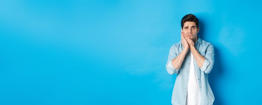 Portrait of man with toothache grimacing from pain and touching cheek, standing against blue background.