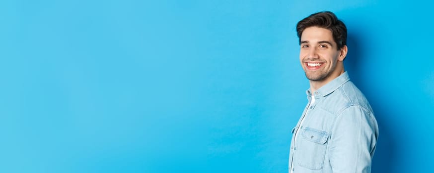 Handsome and confident guy turn head at camera, smiling happy, standing over blue background.