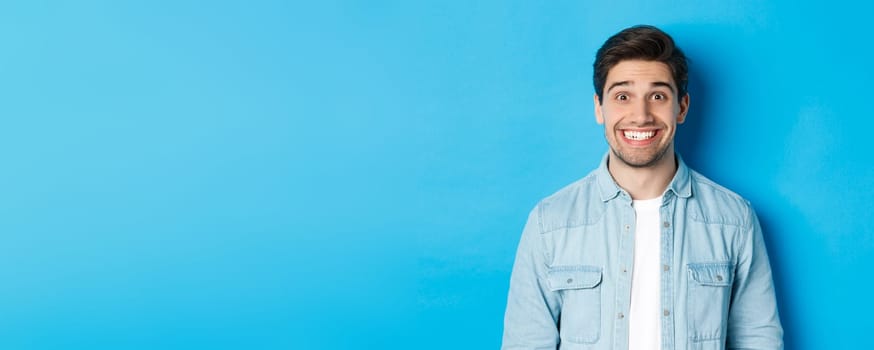 Close-up of smiling excited man with beard, looking amused at advertisement, standing against blue background.