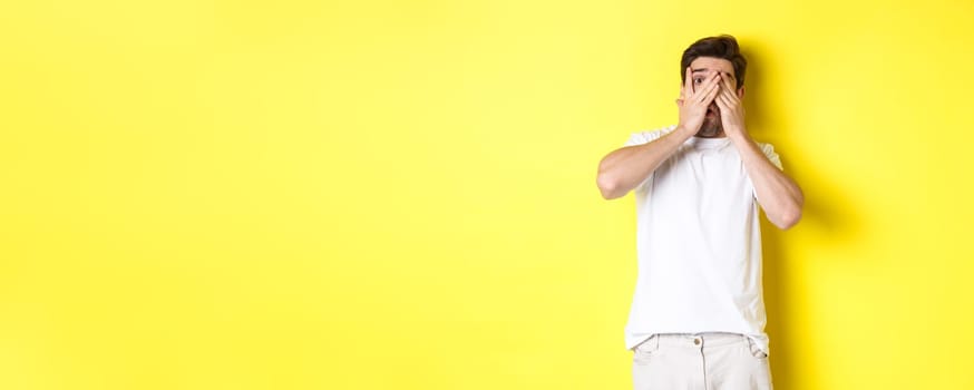 Embarrassed guy shut eyes but peeking through fingers at something awkward, standing over yellow background.