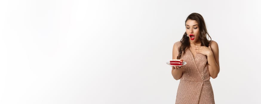 Party and celebration concept. Image of amazed slim female model in luxury dress, looking at piece of cake excited, standing over white background.