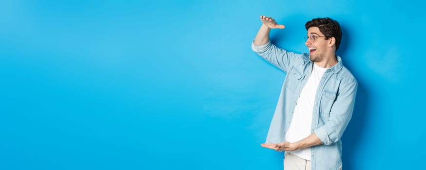 Excited handsome man showing big size object and looking amazed, standing over blue background.