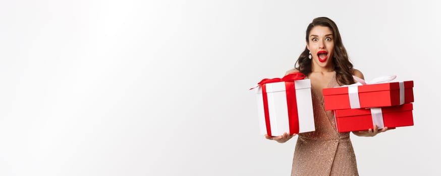 Holidays, celebration concept. Excited and surprised woman holding Christmas gifts and smiling amazed, wearing glamour dress, standing over white background.