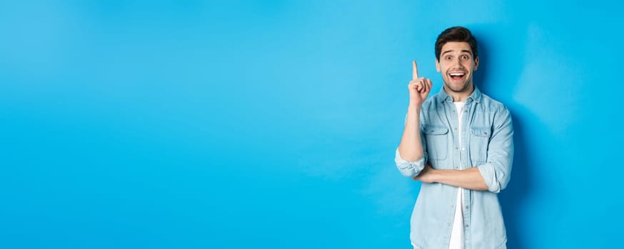 Image of handsome adult man having an idea, raising finger and smiling excited, found solution, standing against blue background.