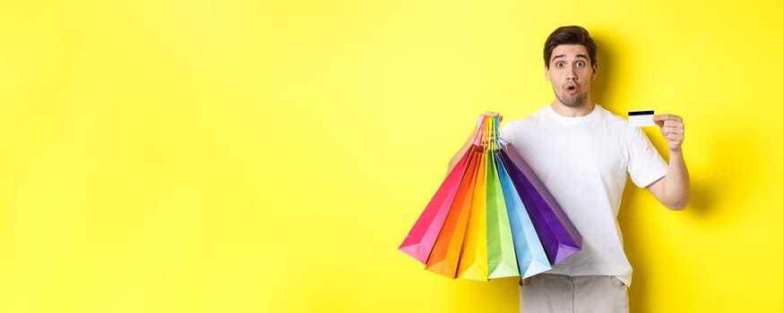 Excited man shopping on black friday, holding paper bags and credit card, standing against yellow background.