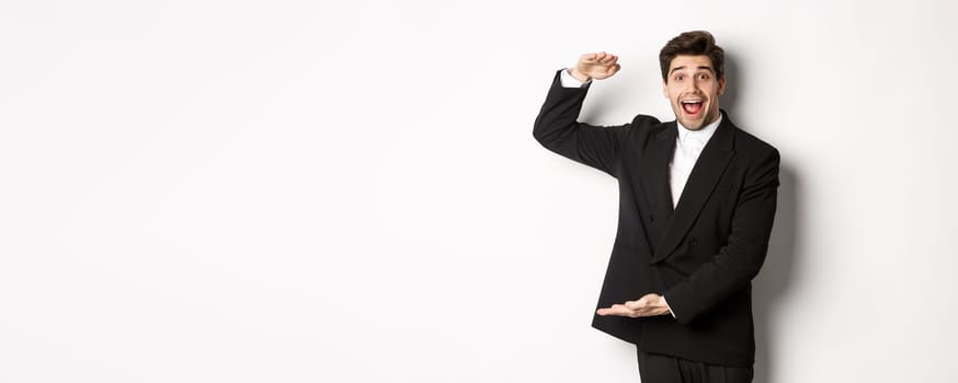 Portrait of excited handsome man in suit, shaping big object on copy space and smiling amazed, holding something, standing over white background.