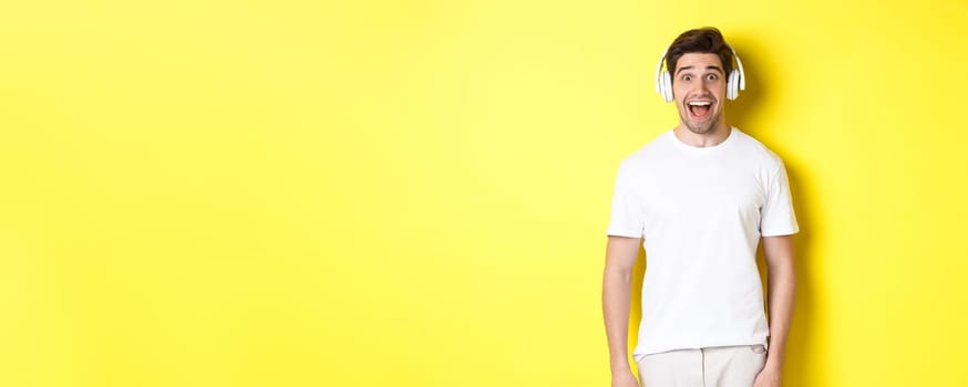 Man in headphones looking surprised, standing against yellow background in white outfit.