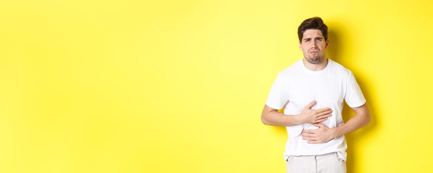 Man having stomach ache, grimacing from pain and touching belly, standing over yellow background.
