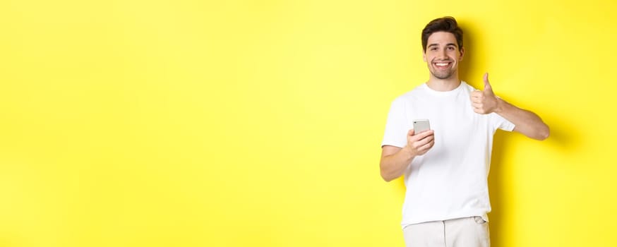 Happy satisfied man holding smartphone, showing thumb up in approval, recommend something online, standing over yellow background.