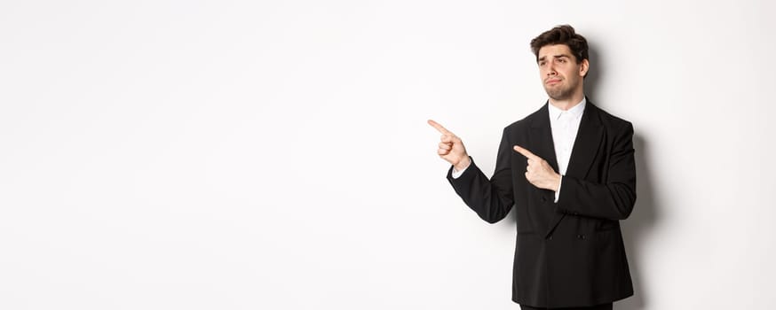 Image of upset and disappointed handsome guy in formal suit, pointing and looking left with sad face, standing over white background.