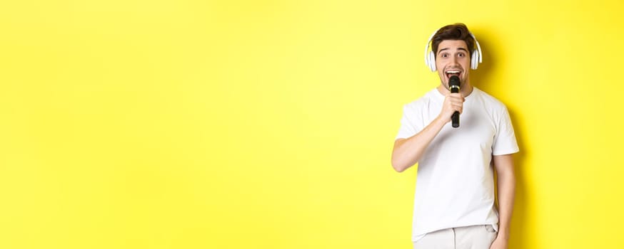 Man in headphones holding microphone, singing karaoke song, standing over yellow background in white clothes.