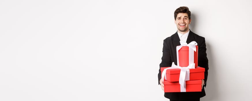 Image of handsome guy in black suit, holding gifts for christmas holidays, standing against white background.