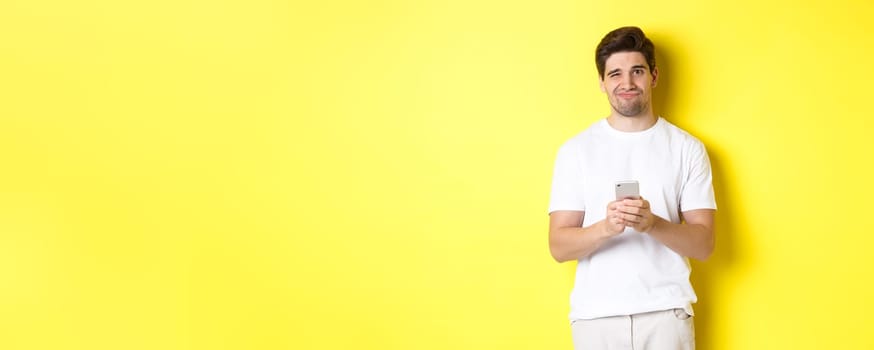 Displeased and reluctant man grimacing, being unamused by message on smartphone, standing over yellow background.