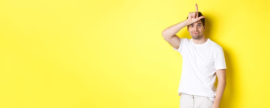 Awkward guy showing loser sign on forehead, looking sad and gloomy, standing in white t-shirt against yellow background.