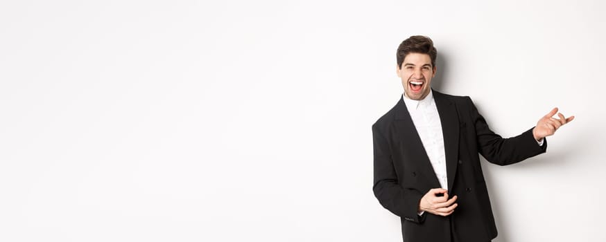 Portrait of happy man dancing at party, playing on invisible guitar and laughing, standing in black suit against white background.