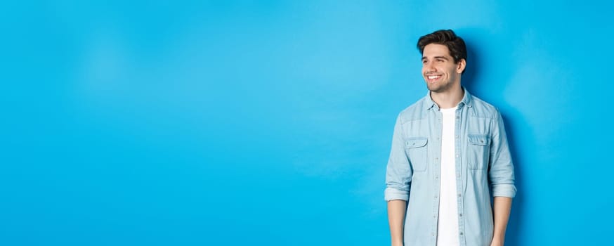 Handsome smiling adult man in casual outfit, smiling and looking left at promo offer, standing against blue background.