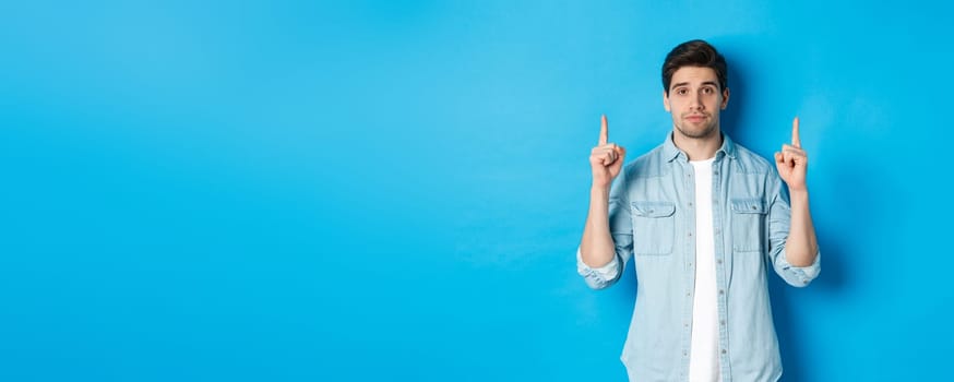 Image of calm handsome man showing you promo offer, pointing fingers up at copy space, standing against blue background.