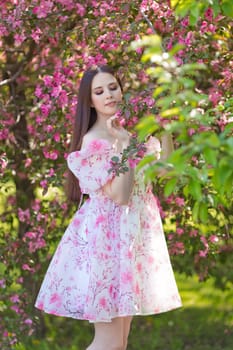 A romantic girl in a light pink dress standing near pink blooming trees, holding a blooming branch, in the garden. Vertical. Close up. Copy space