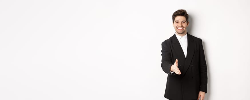 Portrait of handsome businessman in black suit, extending hand for handshake, greet business partners and smiling, welcome to company, standing over white background.