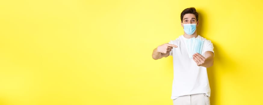 Concept of covid-19, quarantine and preventive measures. Young caucasian man giving medical masks for you, standing against yellow background.