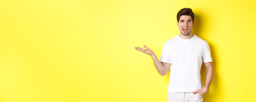 Confused and shocked man complaining, raising one hand and looking bothered, standing near yellow copy space.