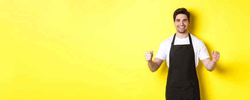 Friendly coffee shop waiter standing with raised hands, place for your sign or logo, standing over yellow background.