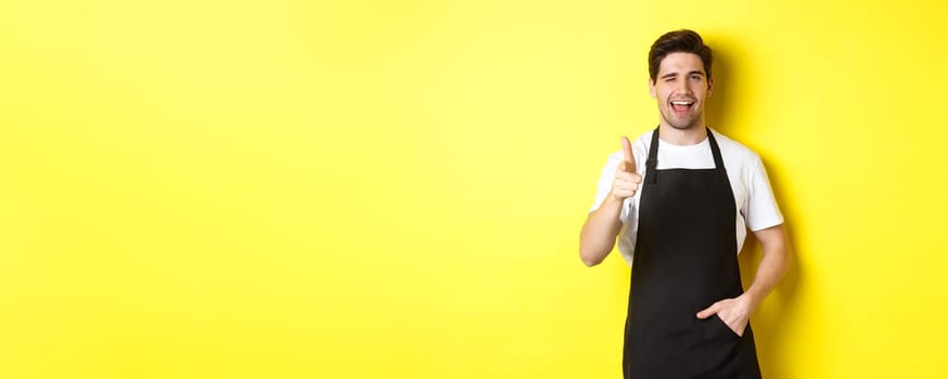 Handsome barista in black apron welcome guests to coffee shop, pointing finger gun and winking, standing against yellow background.