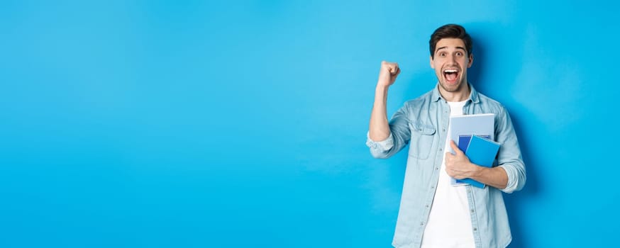 Cheerful guy holding notebooks and celebrating, making fist pump and shouting yes with excitement, standing over blue background.