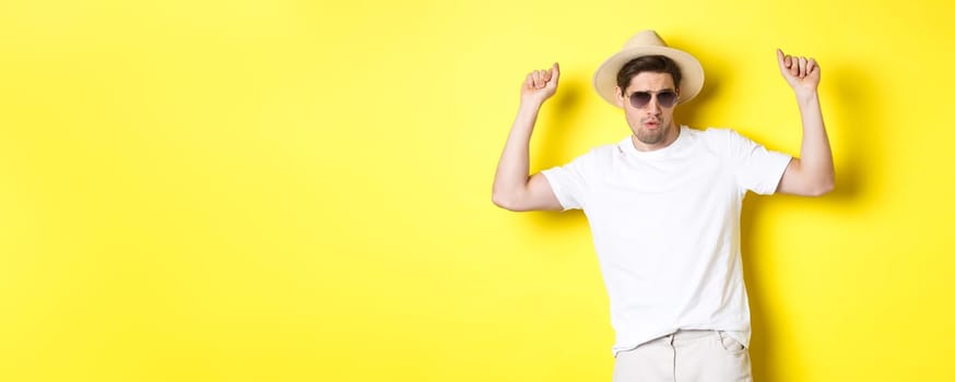Tourism, travelling and holidays concept. Man tourist enjoying vacation, dancing in straw hat and sunglasses, posing against yellow background.