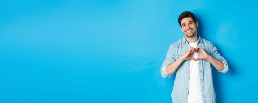 Handsome man smiling, showing heart gesture and looking at camera, saying I love you, standing against blue background.