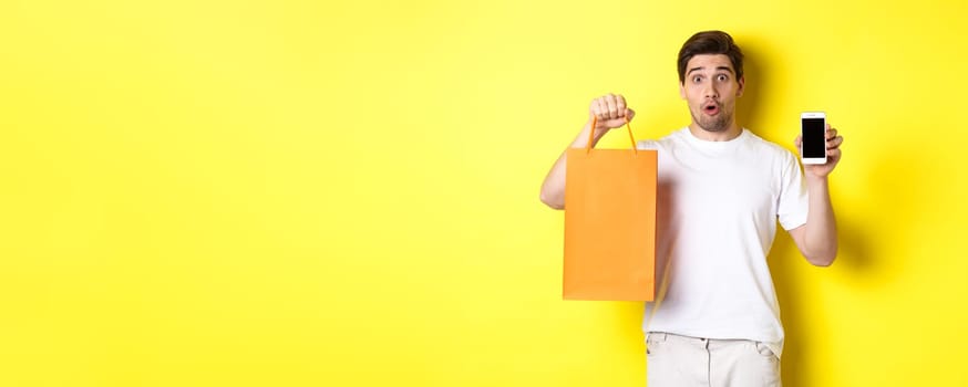 Surprised man showing mobile screen and shopping bag, standing against yellow background. Copy space