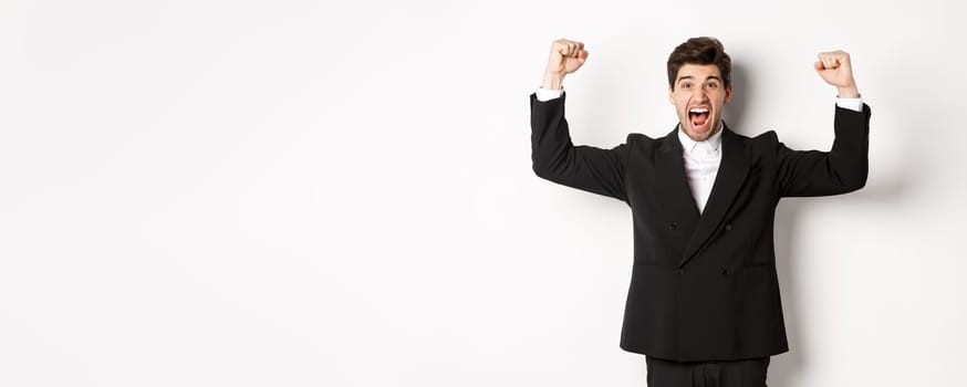 Successful handsome businessman triumphing, raising hands up and shouting yes, rejoicing over achievement, standing against white background.