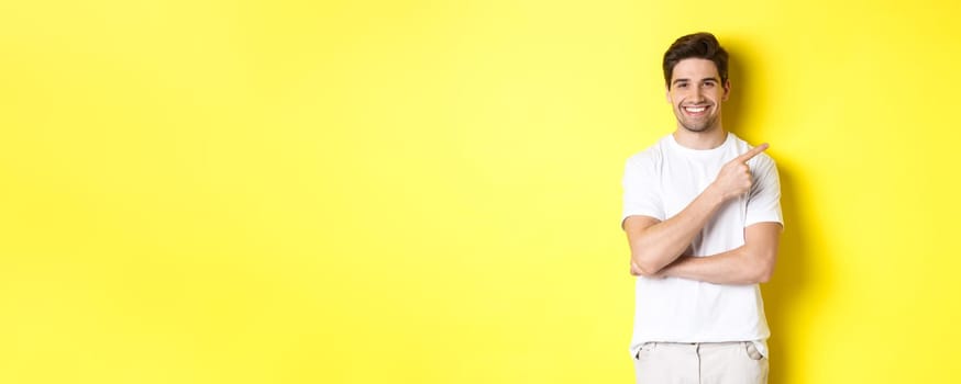 Image of attractive young man pointing finger right at copy space, showing banner or promo offer, standing over yellow background.
