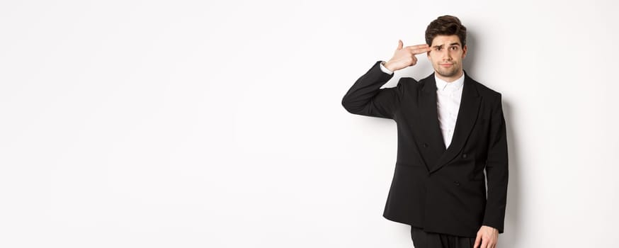 Portrait of distressed and troubled businessman in suit, pointing finger gun at head and looking bothered, shooting himself, standing against white background.