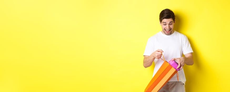 Concept of holidays and celebration. Young man looking surprised as take out gift from shopping bag, standing over yellow background.