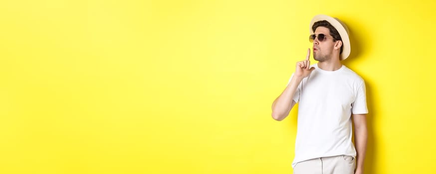 Cool young male tourist blowing at finger gun and looking confident, standing against yellow background. Vacation and lifestyle concept.