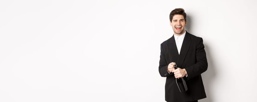 Image of handsome, confident man in black suit, celebrating holiday, open a bottle of champagne and partying, standing joyful against white background.