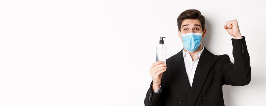 Concept of covid-19, business and social distancing. Close-up of happy man in trendy suit and medical mask, cheering and raising hand up, showing hand sanitizer, white background.