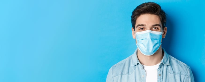 Concept of covid-19, pandemic and quarantine. Close-up of happy guy in medical mask looking at camera, standing over blue background.